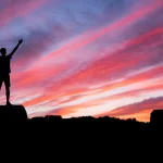 Outlines of a man that uplevels his life now,standing on a rock before a purple, pink and orange sunset.